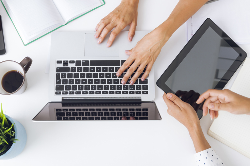 above view of 1 pair of hands typing on a laptop and another holding an ipad