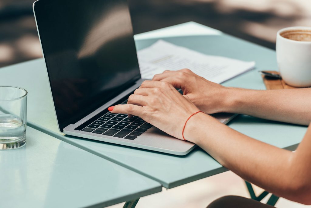 woman's hands typing on a laptop on a blue patio table with a cup of coffee and a stack of papers nearby