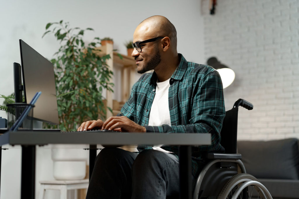 man in a wheelchair wearing a plaid shirt working on a computer at a desk